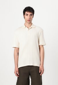 Ung mand med mørkt krøllet hår iført en cremefarvet ribbet kortærmet polo trøje og mørke shorts, stående mod en plain hvid baggrund.