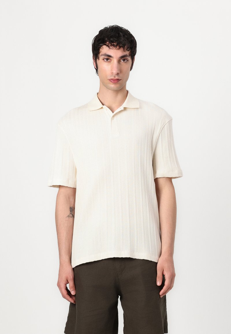 Ung mand med mørkt krøllet hår iført en cremefarvet ribbet kortærmet polo trøje og mørke shorts, stående mod en plain hvid baggrund.
