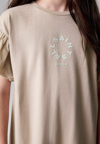 Beige kortärmad t-shirt med rund halsringning och samlad axeldetalj. Har en cirkulär logotyp med texten "SAINT LAURENT" och "LONDON".