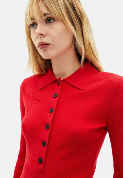 Rød ribstrikket kjole med krave, med knapper foran med seks sorte knapper og en figursyet silhuet. Blød tekstur og glat finish.