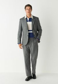 Grijze geblokte pak met een wit en blauw gestreept poloshirt. Schoenen zijn zwart. Op maat gemaakt, gladde textuur, notch-lapels, ontspannen broek.