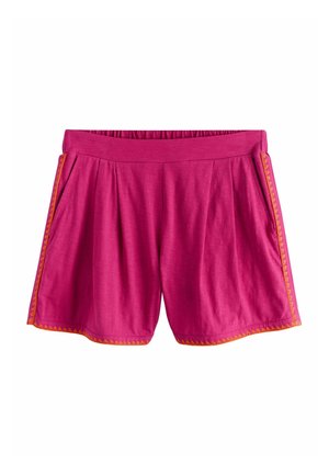 Junge Frau mit langen dunklen Haaren, die ein rotes V-Ausschnitt-Top und passende Shorts mit orangefarbener Borte trägt, steht an einem Sandstrand am Ozean.