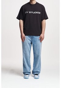 Svart bomulls-t-shirt med "2Y STUDIOS" tryckt i vitt, kombinerad med ljusblå jeans och svartvita sneakers med blå detaljer.