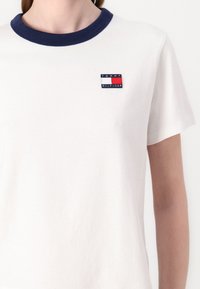 Hvid bomulds T-shirt med en marineblå ribbet krave og et lille, rektangulært logo patch med brandnavnet og farvede striber.