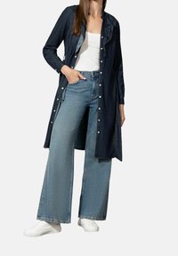 Denim-paitakleid mit Rüschekragen, dunkelblauem Stoff, weißen Knöpfen, kombiniert mit hellblauen, weiten Jeans und weißen Sneakers.