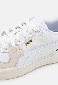 Baskets en cuir blanc avec des accents en daim beige, bout rond, lacets plats et logo doré. Présente une surface texturée et des coutures contrastées.
