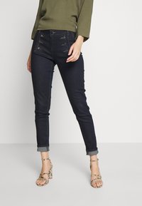 Mörkblå denimjeans med slim passform, med sex dekorativa knappar fram. Uppvikt fåll och kombinerade med sandaler med klack och remmar.