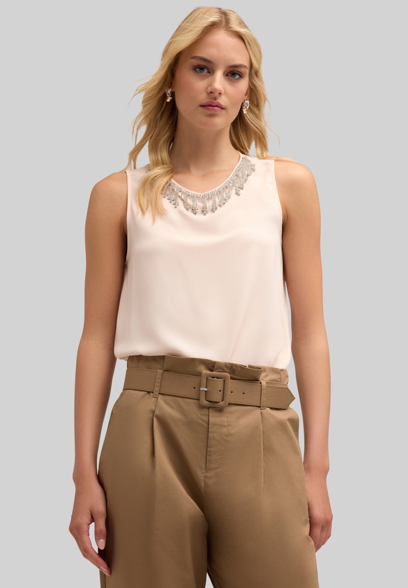 Ärmlös ljusrosa topp med detalj av stenar vid halsen, i kombination med högmidjade beige byxor med matchande bälte och veck.