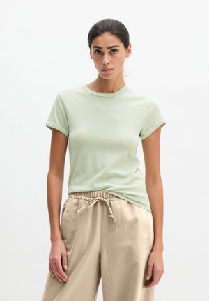 Frau mit zurückgebundenen dunklen Haaren trägt ein hellgrünes T-Shirt mit kurzen Ärmeln und beige Kordelzughose, steht vor einem einfachen weißen Hintergrund.