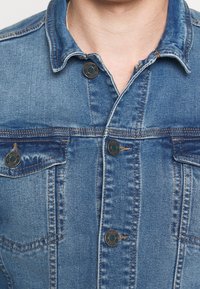 Jeansjacka, blåtvätt, med knapplist fram, två bröstfickor och klassisk krage. Sliten textur och synliga sömnadsdetaljer.