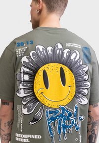 Camiseta de algodón verde oliva con un gran gráfico de una cara sonriente amarilla con pétalos negros y texto goteando azul, con diseños impresos adicionales.