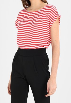 Femme portant un haut à manches courtes rayé horizontalement rouge et blanc, rentré dans un pantalon noir taille haute, sur un fond uni.