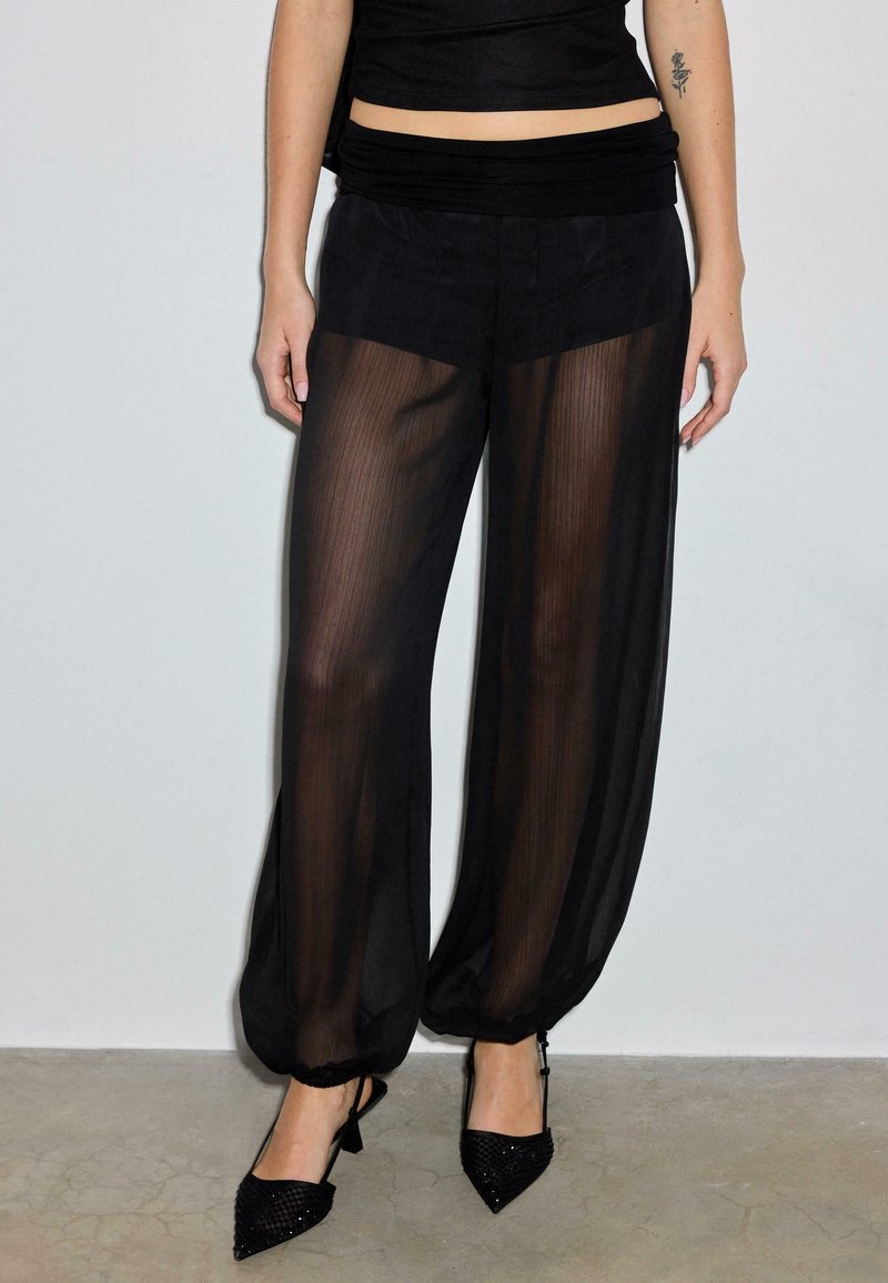 Schwarze, weit geschnittene Hose aus transparentem Stoff mit strukturierter Oberfläche, gerafftem elastischem Bund und elastischen Bündchen. Dazu werden spitze schwarze Absatzschuhe getragen.