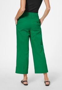 Groene wijde broek met een gladde textuur, voorzien van een achterzak met knoop en een omgeslagen zoom. Gepaard met zwarte sandalen.