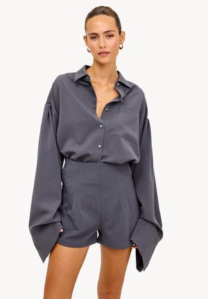 Femme portant une chemise boutonnée gris foncé surdimensionnée avec des manches larges et un short taille haute assorti, debout devant un fond uni.