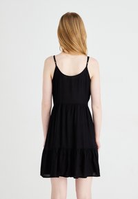 Rochie neagră fără mâneci, cu bretele ajustabile, talie strânsă și tiv învoltat cu volane, realizată dintr-un material ușor. Vedere din spate.