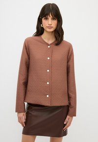 Brun vadderad jacka med rund halsringning, med texturerad tyg och vita knappar, kombinerad med en mörkbrun läderkjol.