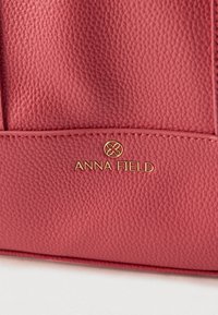 Közelkép texturált piros bőr kézitáskáról, aranyszínű Anna Field logóval és varrott varratokkal.