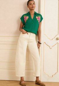 Blusa corta de manga corta en color verde con bordado floral, combinada con pantalones blancos de pierna ancha y cintura alta, y zapatos planos marrones. Fondo de suelo de madera natural.