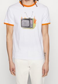 Person iført en hvid t-shirt med orange krave og ærmekant, der har et retro fjernsyn i brand grafik i midten af brystet.
