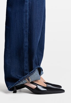 Czarne skórzane buty na szpic z wycięciami po bokach i niskim obcasem typu kitten, zestawione z podwiniętymi ciemnymi jeansami. Oba elementy są sfotografowane z bliska i wyraźnie widoczne.