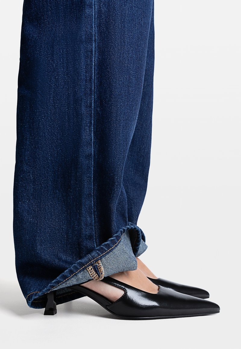 Chaussures en cuir noir à bouts pointus avec des côtés découpés et un petit talon, associées à un jean en denim foncé retroussé. Les deux sont en gros plan et bien définis.