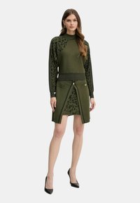 Donna che indossa un set composto da maglione verde e gonna con dettagli in stampa leopardata nera e tacchi alti neri, in piedi su uno sfondo bianco.