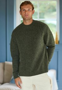Un pull en maille vert foncé avec un col rond et des poignets côtelés, associé à un pantalon beige clair, le tout dans un décor intérieur doucement éclairé.