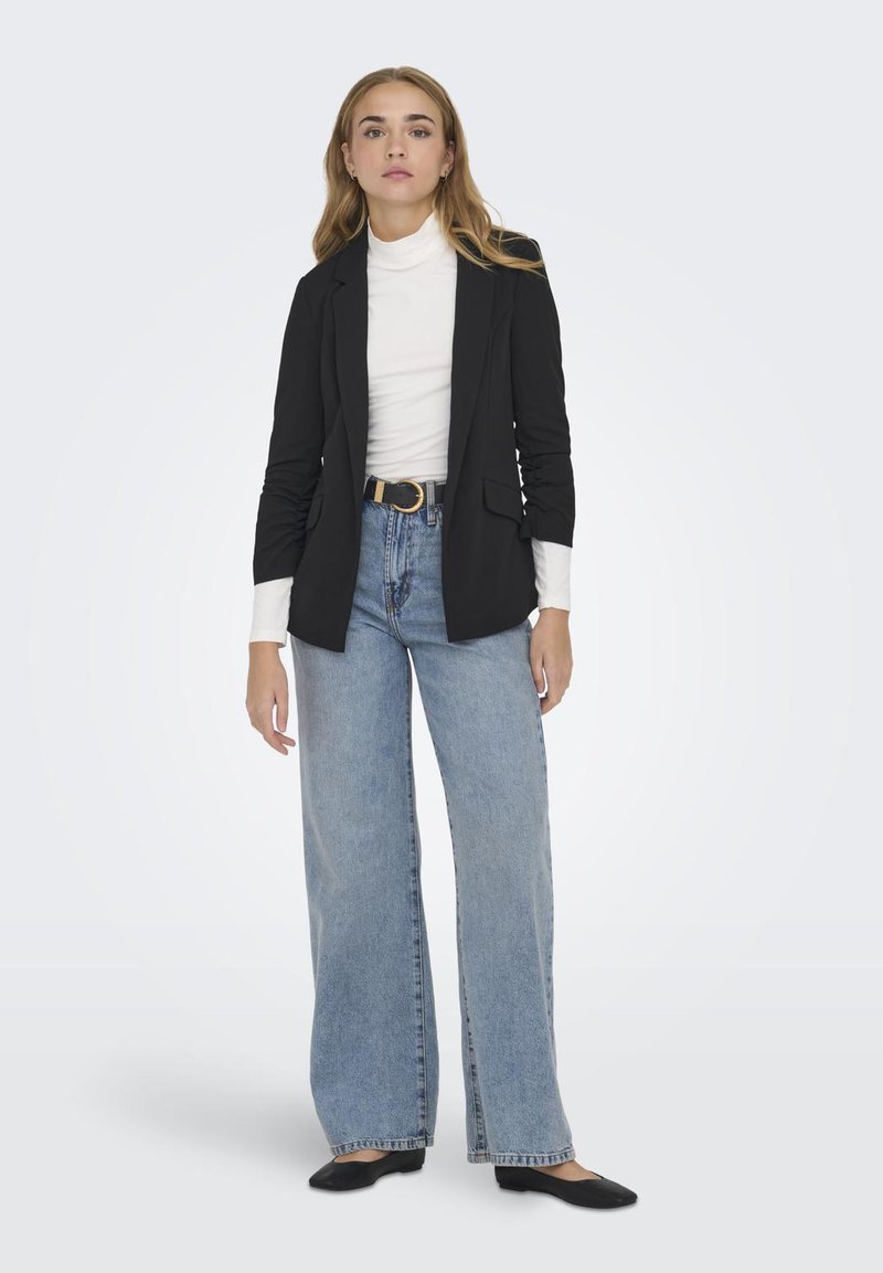 Zwarte blazer met een getailleerde pasvorm, gecombineerd met een witte turtleneck en lichtblauwe hoge taille wijde jeans, aangekleed met een zwarte flat.