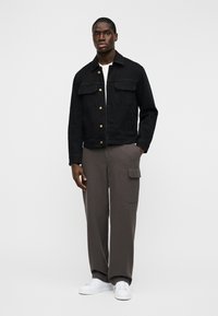 Μαύρο τζιν τζάκετ με χρυσά κουμπιά, δύο τσέπες στο στήθος, φορεμένο πάνω από λευκό πουκάμισο και γκρι παντελόνια με πλαϊνή τσέπη cargo και λευκά παπούτσια.