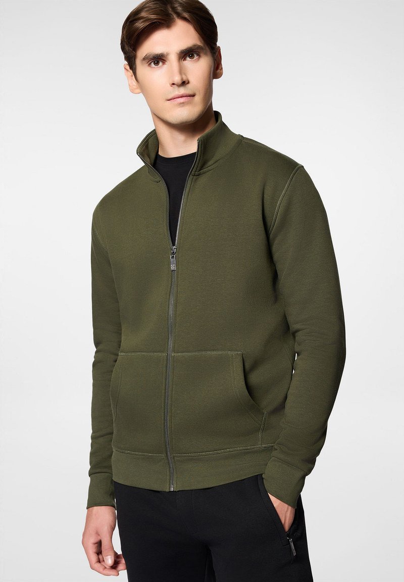 Oliwkowa bluza z zamkiem błyskawicznym i wysokim kołnierzem, z dwoma przednimi kieszeniami, ściągaczami na rękawach oraz prostym dołem, wykonana z teksturowanej tkaniny bawełnianej w składzie.