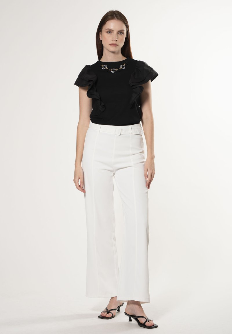 Blusa negra con mangas acampanadas y escote bordado, combinada con pantalones blancos de cintura alta, finalizada con un cinturón elegante y tacones negros.