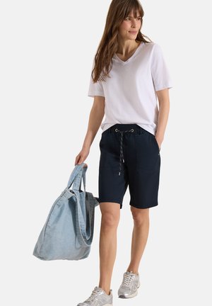 Kvinde iført hvid V-hals T-shirt og marineblå shorts, bærende en stor lyseblå tote-taske og gående i sølvfarvede sneakers.