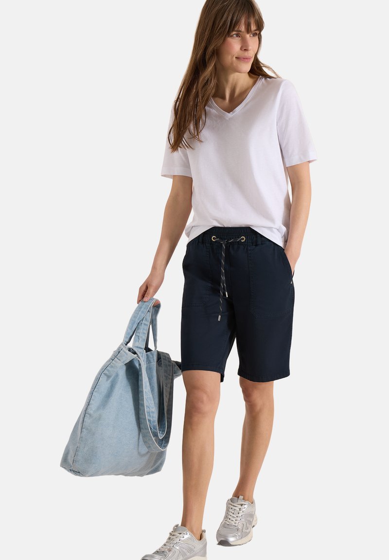 Mujer con camiseta blanca de cuello en V y pantalones cortos azul marino, llevando un gran bolso tote azul claro y caminando con zapatillas plateadas.