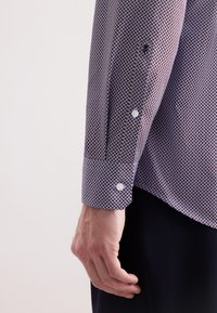 Camicia con bottoni in un motivo geometrico marrone scuro e azzurro chiaro, con polsini dotati di due bottoni bianchi. Tessuto dalla superficie liscia.