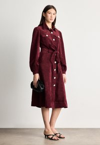 Burgundi kordbársony ruha, galléros nyakkivágással, hosszú ujjal, elülső gombokkal és szövet övvel. Fekete kézitáskával és szandállal kombinálva.