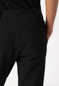 Crne hlače glatke teksture, prilagođenog dizajna, s stražnjim džepom. Pojas je elegantan, a tkanina djeluje lagano.