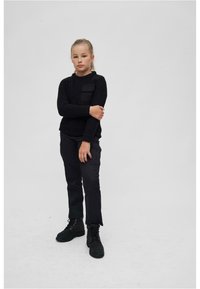 Maglione nero a maglia con taschino sul petto, abbinato a pantaloni cargo neri e stivaletti neri con lacci. Il modello è in piedi di fronte a uno sfondo chiaro.