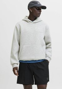 Ein schwarzer Mann trägt einen hellgrauen Hoodie, schwarze Shorts, eine marineblaue Kappe und kleine Creolen, während er seitlich gegen einen schlichten Hintergrund schaut.