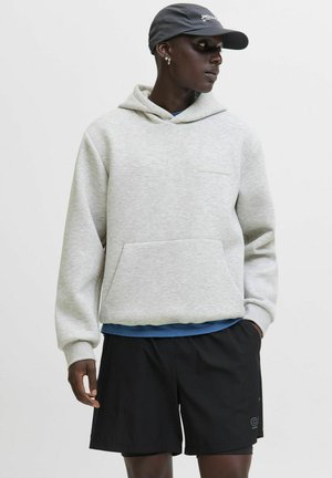 Ein schwarzer Mann trägt einen hellgrauen Hoodie, schwarze Shorts, eine marineblaue Kappe und kleine Creolen, während er seitlich gegen einen schlichten Hintergrund schaut.