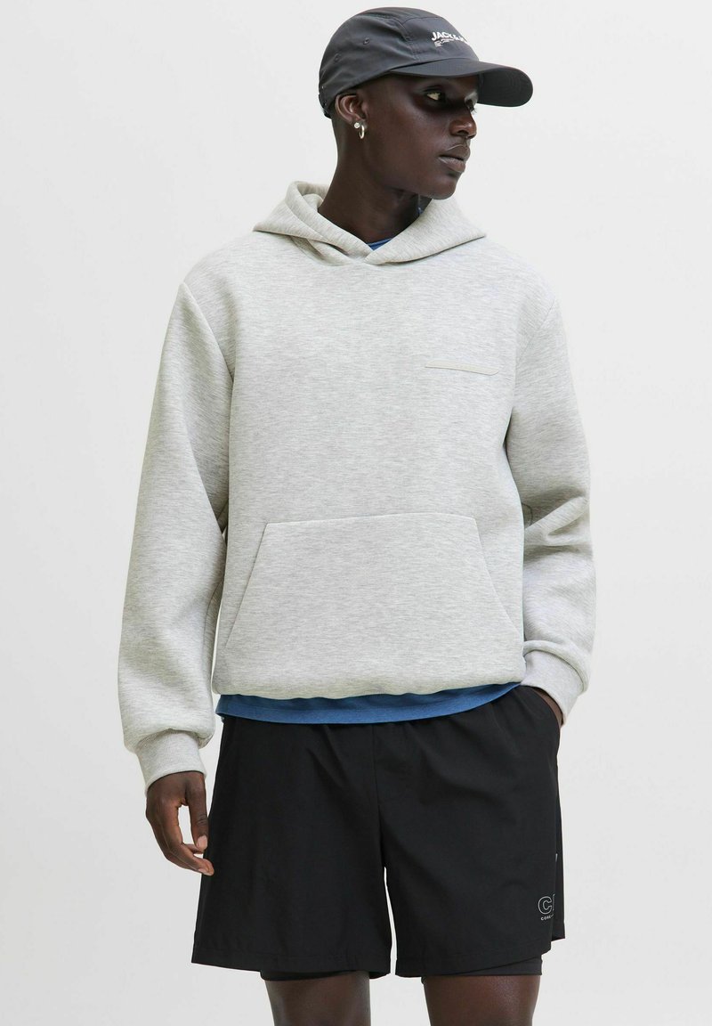 Ein schwarzer Mann trägt einen hellgrauen Hoodie, schwarze Shorts, eine marineblaue Kappe und kleine Creolen, während er seitlich gegen einen schlichten Hintergrund schaut.
