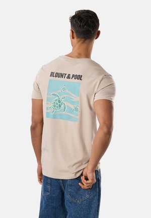 Mand iført beige T-shirt med blå grafik af tre svømmende skildpadder og teksten "BLOUNT & POOL" på ryggen, kombineret med blå denimjeans.