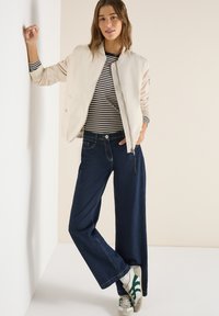 Jeune femme portant une veste bomber beige, un chemisier à rayures, un jean large foncé et des baskets blanches, appuyée contre un mur dans un décor neutre.