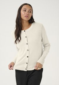 Svetlo bež kardigan iz mehkega materiala, z okroglo izrezom, dolgimi rokavi in temnimi gumbi spredaj. Gladka tekstura.