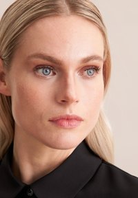 Nahaufnahme eines Gesichts einer Frau mit heller Haut, hellblonden Haaren, hellblauen Augen und neutralen Lippen, die ein schwarzes Hemd mit Kragen trägt.