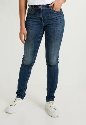 Jeans skinny de color azul oscuro confeccionados en denim. Cuenta con cinco bolsillos, trabillas para el cinturón y un ligero desgastado a lo largo de los muslos. Combinados con zapatillas blancas.