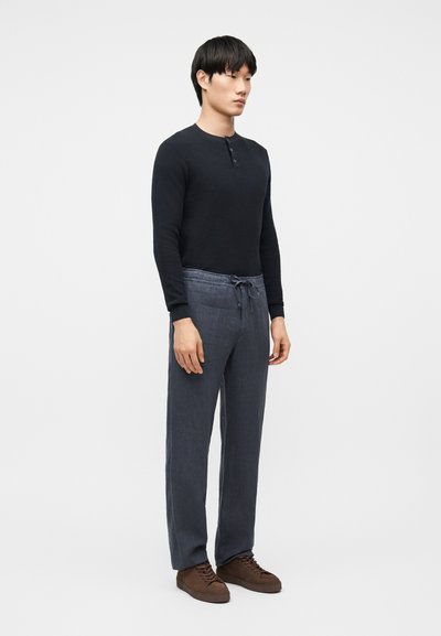 Άνδρας που φορά μαύρο μακρυμάνικο πουκάμισο henley, γκρι παντελόνι με κορδόνι και καφέ παπούτσια με κορδόνια, στέκεται μπροστά σε απλό φωτεινό φόντο.