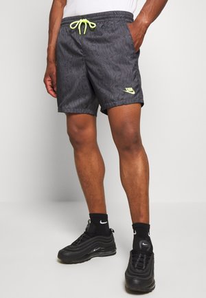Mężczyzna w czarnych szortach z wzorem, z neonowym sznurkiem i logo Nike, czarne skarpetki Nike oraz czarne buty sportowe Nike, stojący na szarym tle.