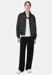 Schwarze Bomberjacke mit gerippten Bündchen und Reißverschluss, kombiniert mit einem weißen Oberteil und weiten schwarzen Hosen. Helle Sneakers runden den Look ab.