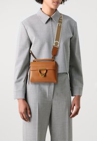 Brun lærcrossbody-veske med strukturert form, gullfarget hardware og justerbar stropp med logo. Mønstret rutete bluse på.
